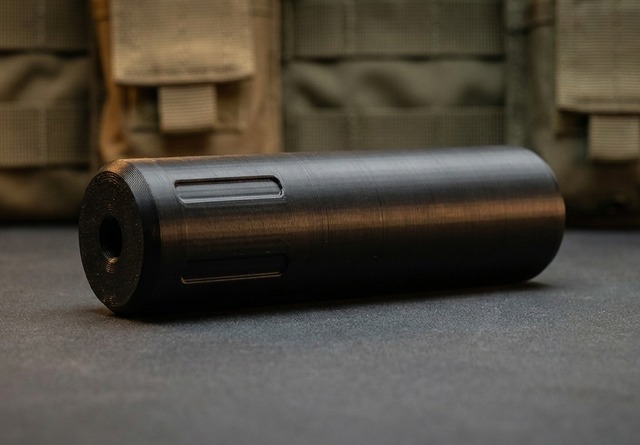 Ballistic Designs Mini Magna SD airsoft moderator lying on a tactical workbench with soft-focus MOLLE pouches behind